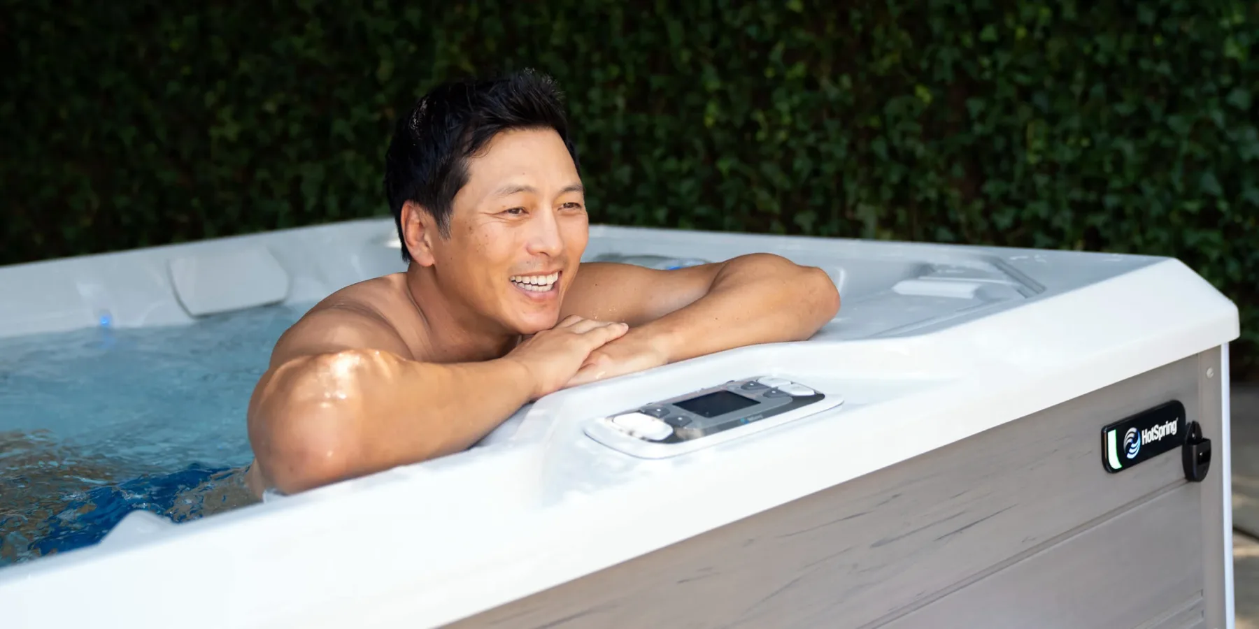 A man resting his arms on the edge of a Hot Spring spa and smiling, a tall green hedge behind him