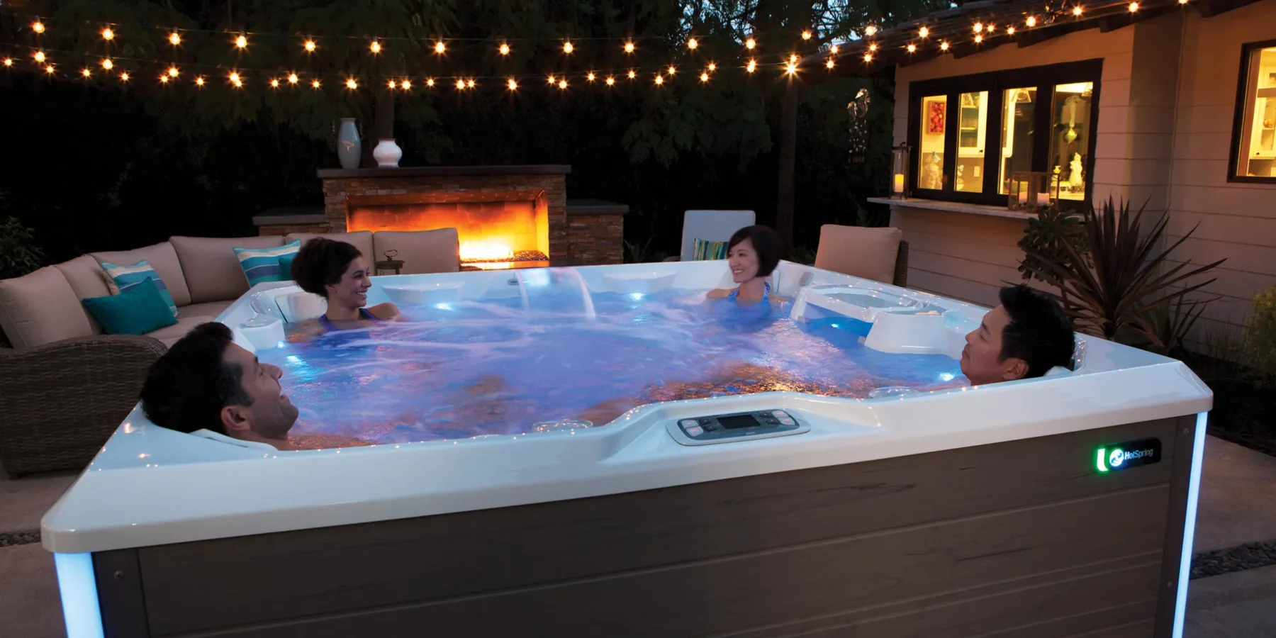 Four friends soaking in a lit Hot Spring spa at dusk, under string lights beside an outdoor fireplace