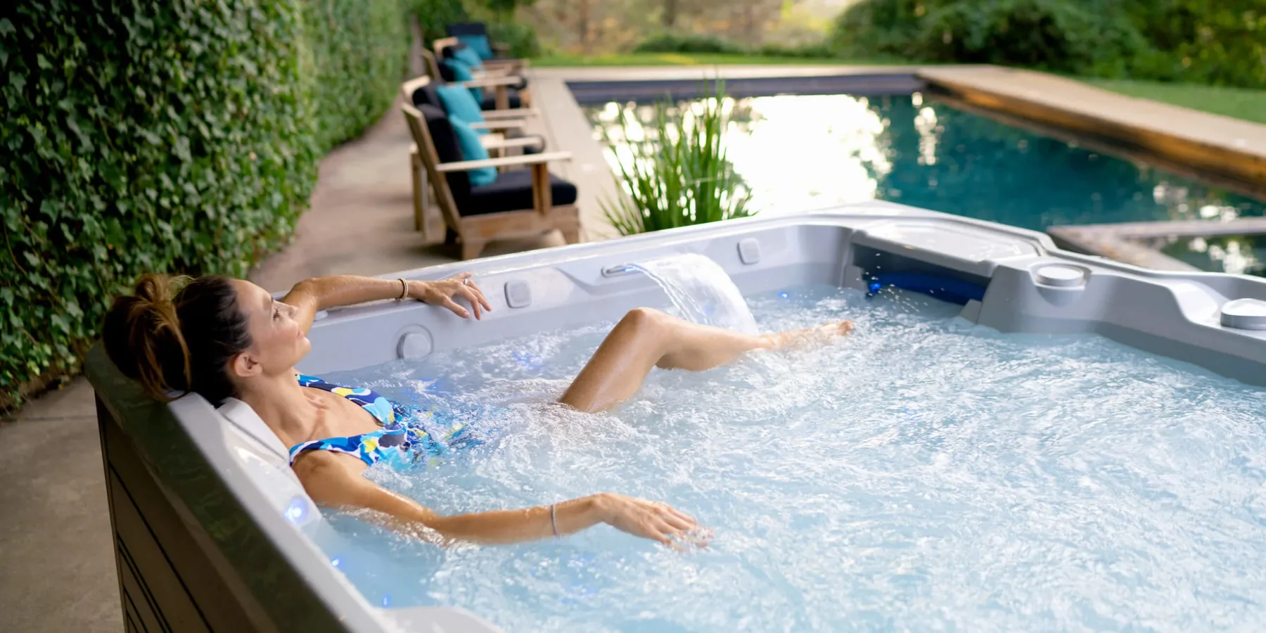 A woman reclining in a Hot Spring spa next to a backyard pool, water pouring from the spa's fountain