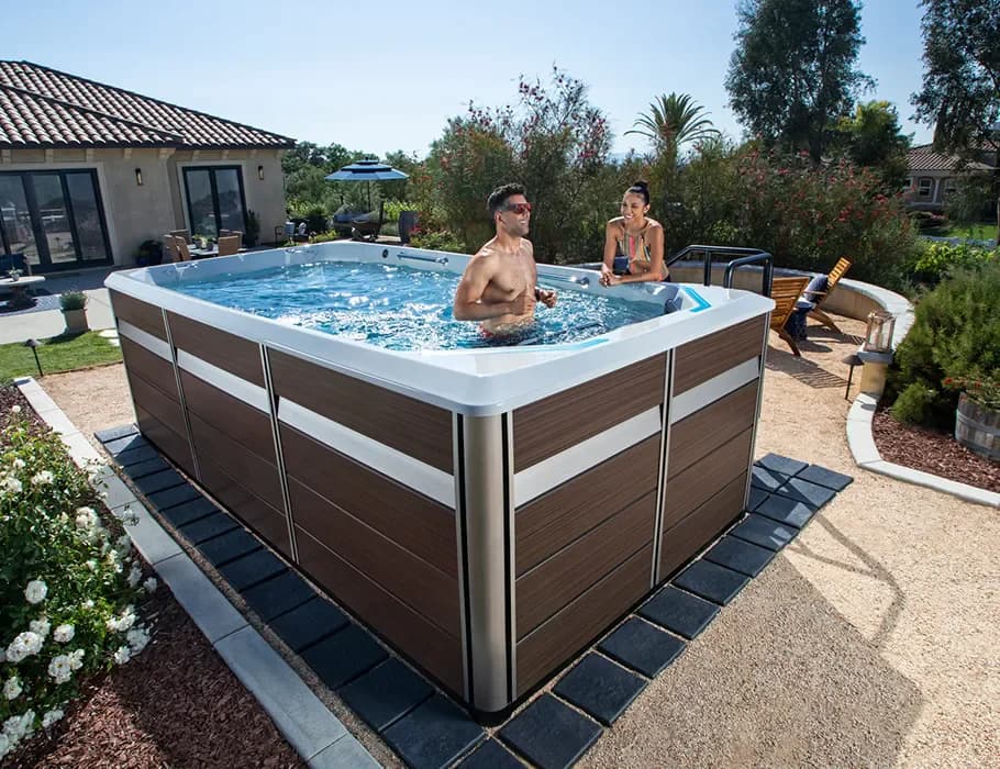 A couple chatting in a swim spa on a sunny patio, one in the water and one leaning on the edge