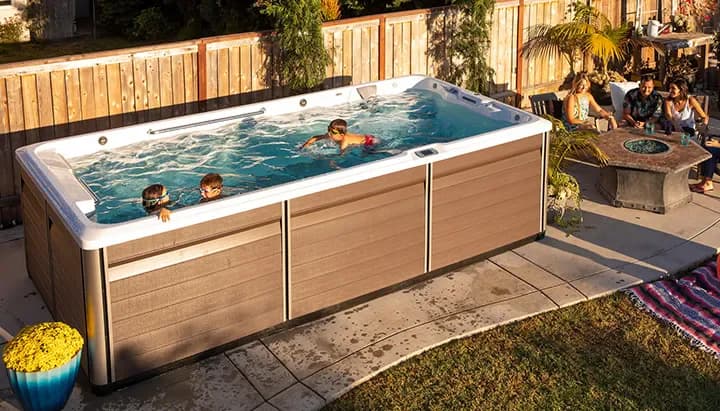 Seen from above, three kids swimming in a backyard swim spa while their parents sit at a fire table beside it